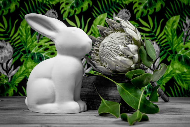 White ceramic bunny sculpture beside a blooming protea, surrounded by lush green leaves.